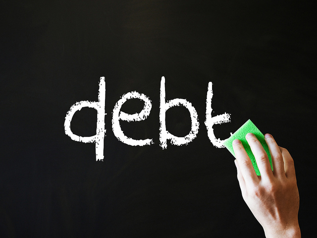 the word "debt" on a chalkboard along with a hand holding an eraser